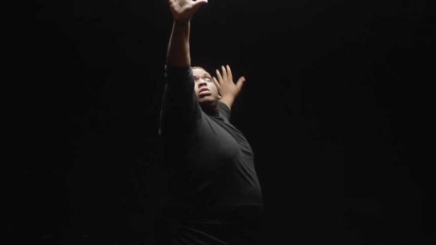 Male dancer wearing dark turtle neck and shorts dances on wooden floor in dark studio lit by overhead lighting