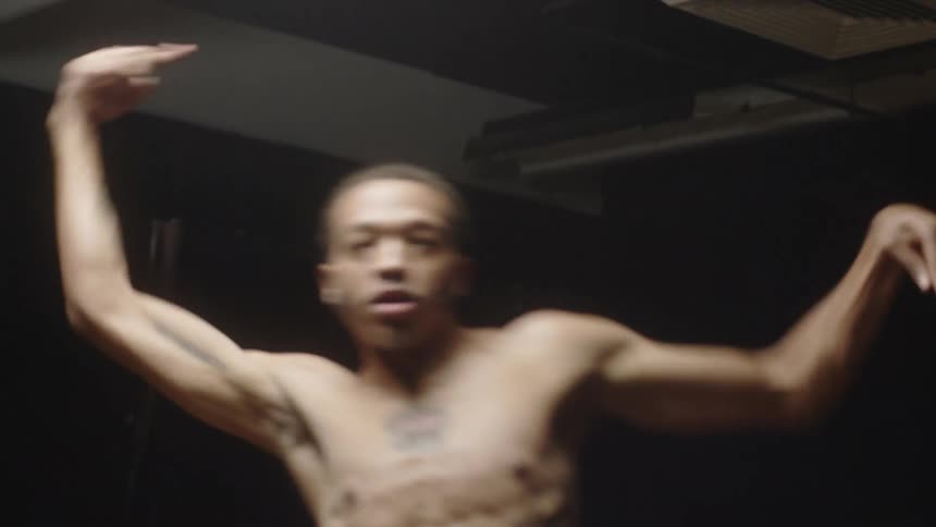 Bare chested male dancer with tattoos dances under bright light in dark room