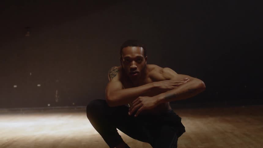 Muscular shirtless man with tattoo black pants and socks dances in spotlight in dark room