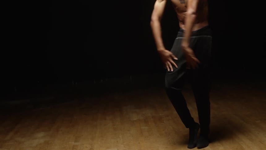Shirtless muscular man with tattoo dances in the spotlight in dark room