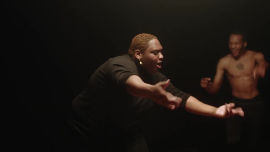 A woman dances under a light in a dark room as two other people watch and dance along with her