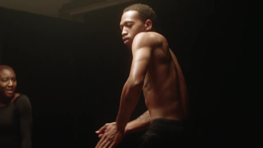 Three women clap and laugh around a topless man as they perform a hip hop dance that ends with them all falling on the floor