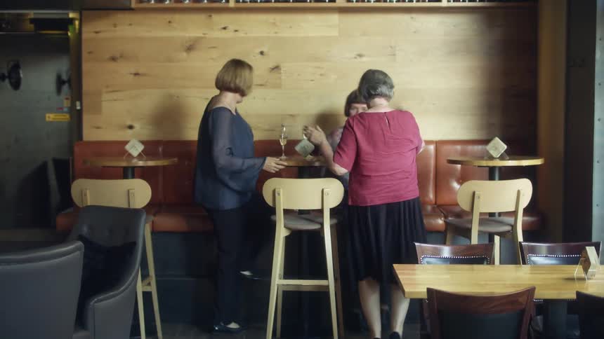 A group of women wearing pants skirt and blouses sits down on couch in pub