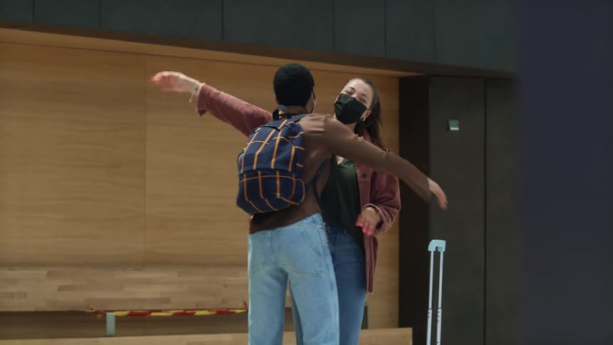 A woman waits at an airport for a friend and greets him with a hug as he walks up with a suitcase