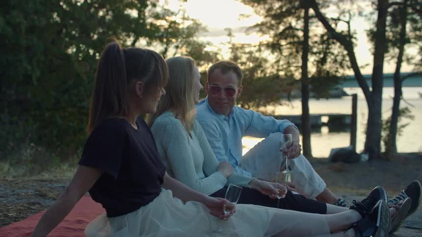Friends talking and laughing while drinking champagne near a lake at sunset