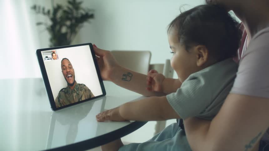 Mother holding toddler boy sits at table making a video call o tablet in a room