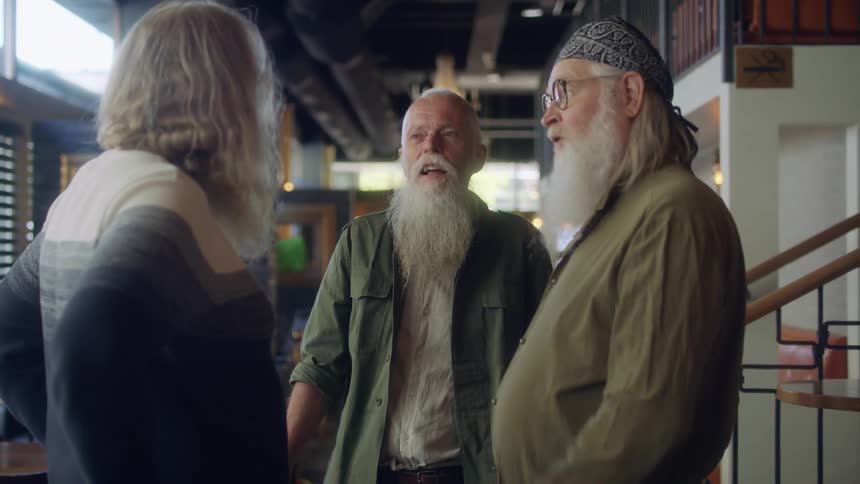 Three friends with long white beards stand near tables in an empty restaurant and chat