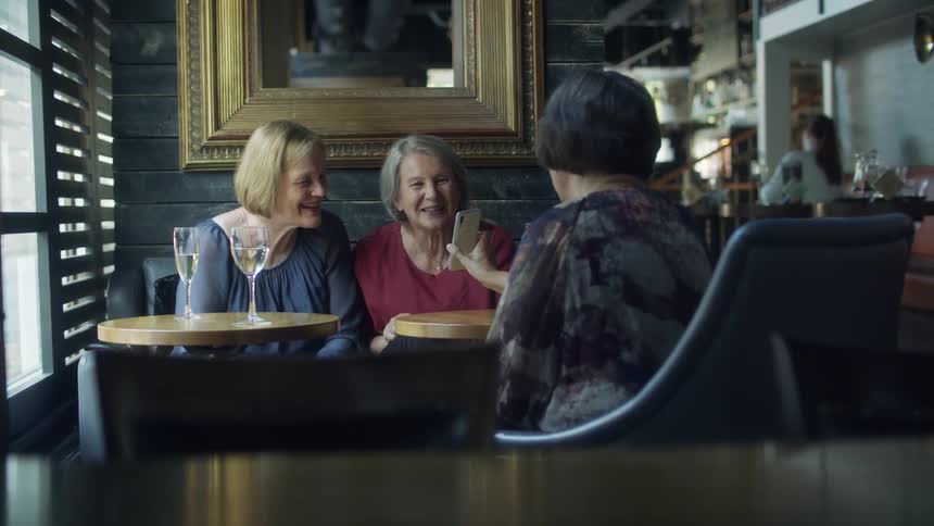 Three women sitting and looking at a smart phone with a wall and window in the background