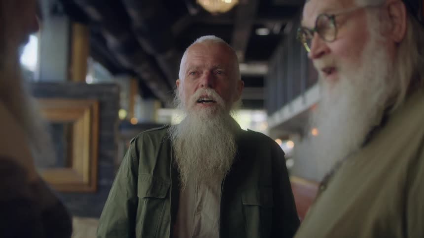 Three older male friends stand in restaurant aisle between tables laughing and talking
