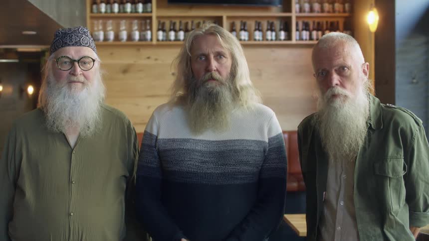 A group of long bearded men stare into the camera while standing side by side indoors