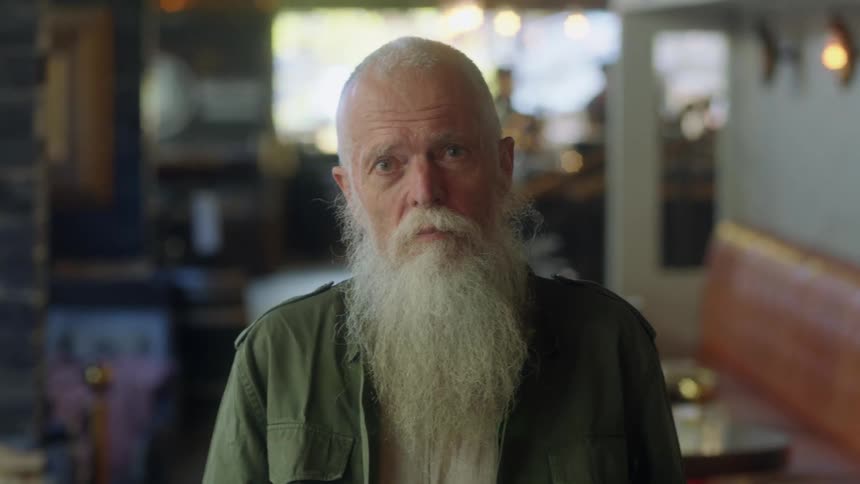 Elderly white haired man with long beard stands between tables at cafe staring