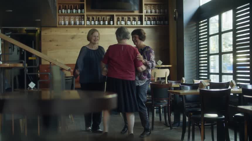 Two women welcome another friend as they hug and go back to sit on their table in a restaurant