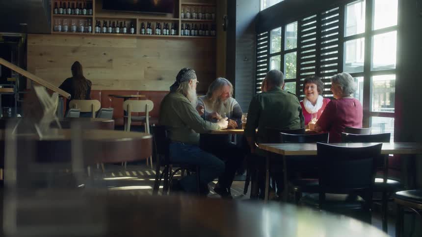 Older friends greet a new arrival at table in restaurant with wines displayed high on wall