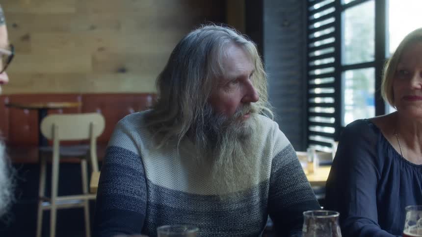 Elderly men with long grey hair and beard and woman sit at table having a conversation in pub