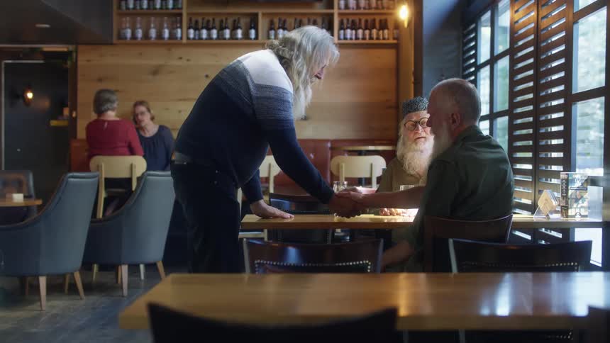 Old friends greet each other with clasped hands and sit to talk at table with alcoholic drinks