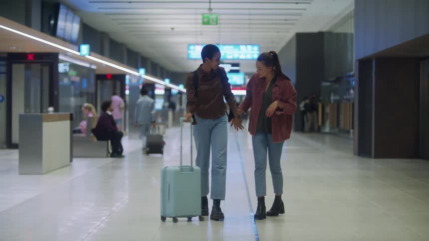 Two women stand and hug each other in the middle of an airport before holding hands and walking together