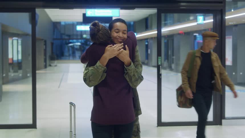 Women hug in greeting near automatic doors at airport terminal as others walk past