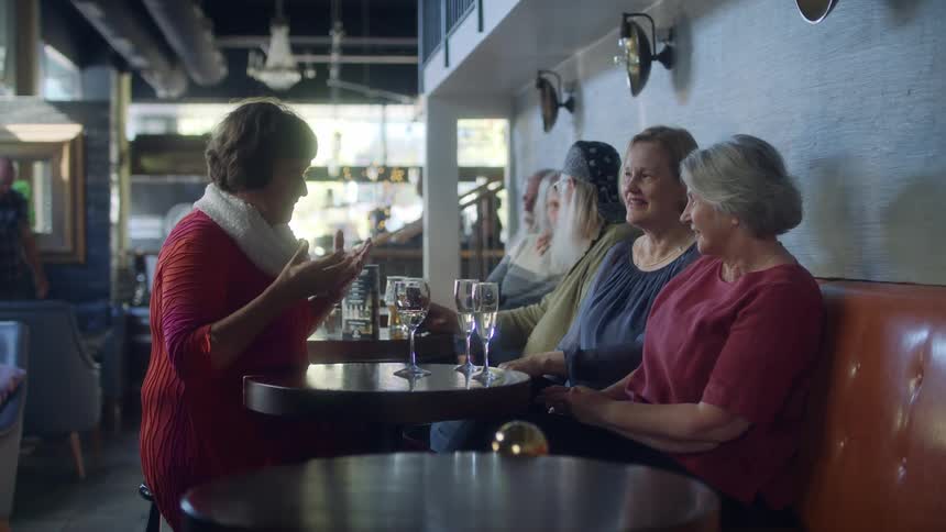 Three women sitting at a table in a restaurant with people a wall and windows in the background