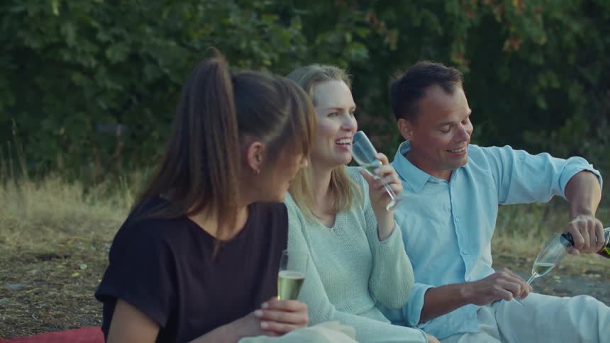 A couple and their friend laugh as they drink champagne in a park