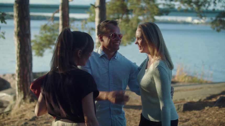 Friends greet each other with hugs and stand talking near trees on shore as vehicle passes along road on opposite shore