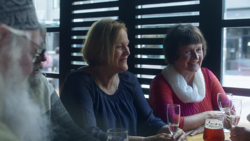 Elderly friends sit at restaurant having drinks as pedestrians walk by window