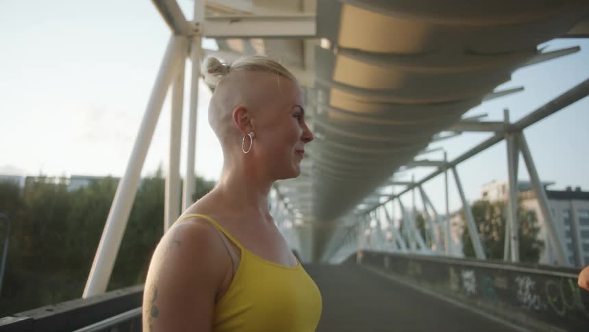 Female dancer with short hair hoop earrings yellow sleeveless shirt and tattoos smiles at her friends on the bridge