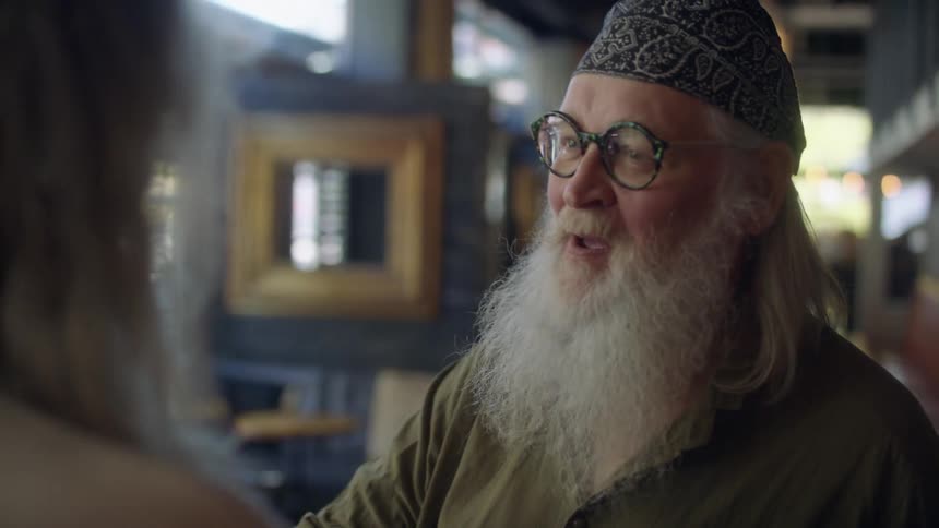 Older man with long beard and his friend greet each other by clasping onto arms at mostly empty restaurant