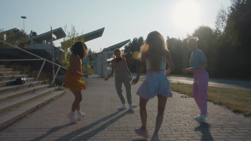 Four women dancing on a sidewalk with trees the sky and clouds in the background