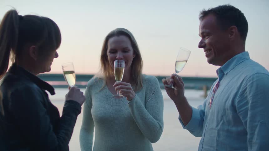 The trio of people smile as they talk and sip on champagne from glasses while they stand outside