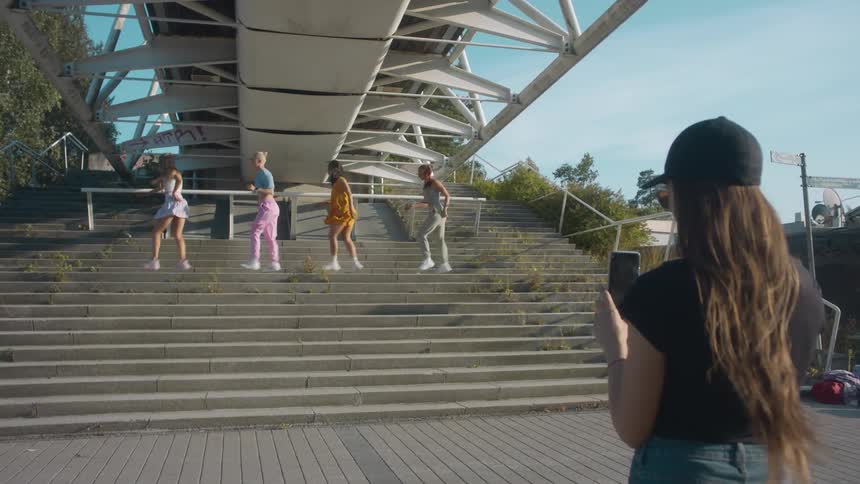 Four young women perform a choreographed dance on steps under a bridge as another friend records it on her phone