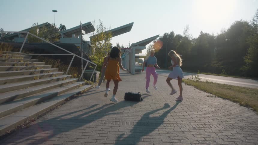 A group of women perform a synchronized dance while surrounding a radio on a walkway outdoors on a sunny day