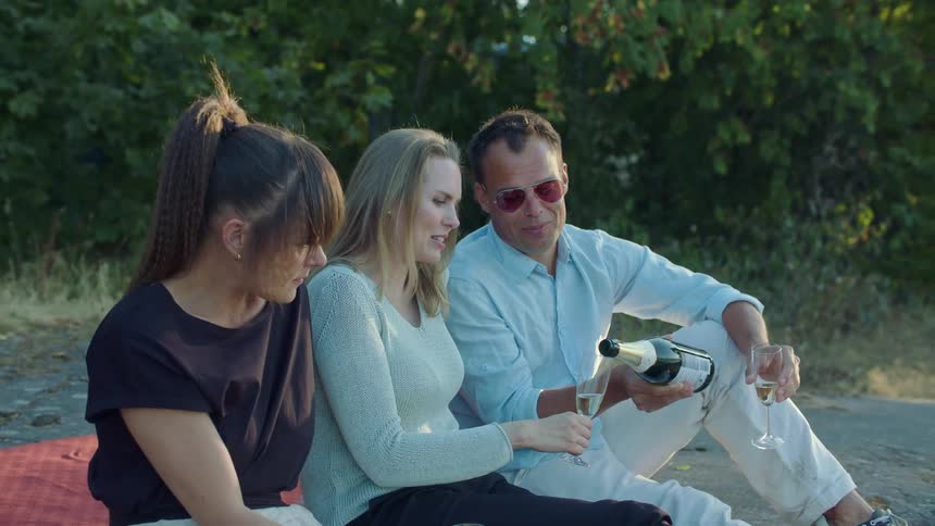 A man pours champagne for two women while sitting on a picnic blanket