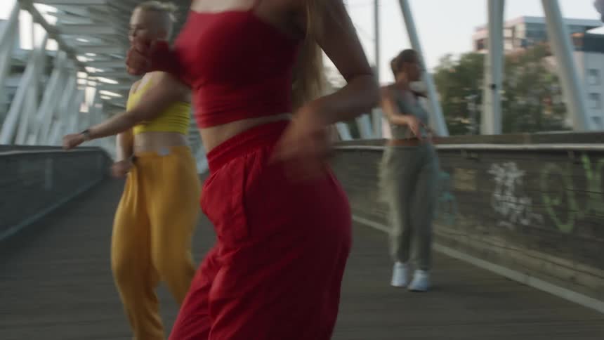 A group of female dancers in brightly colored clothing dance on a pedestrian bridge thats covered in graffiti