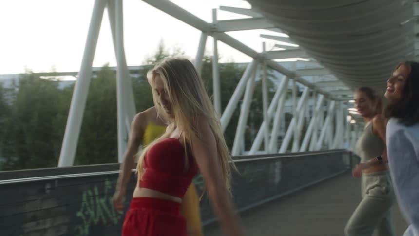 Four women are dancing on a walking bridge in an urban area at dusk