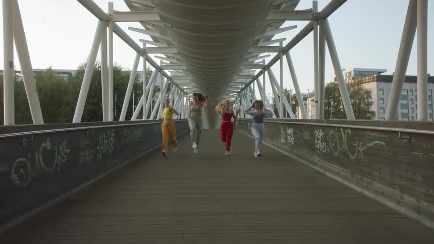 Four female friends run laughing across graffiti covered pedestrian bridge with buildings in background