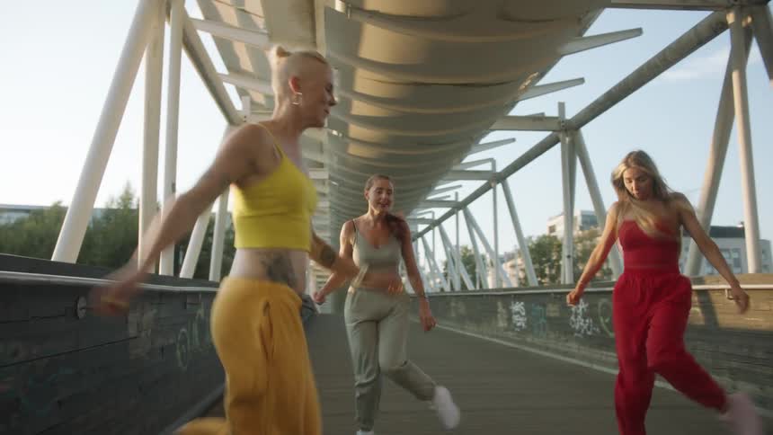 Four female friends dance on covered pedestrian walkway decorated with graffiti with buildings in background