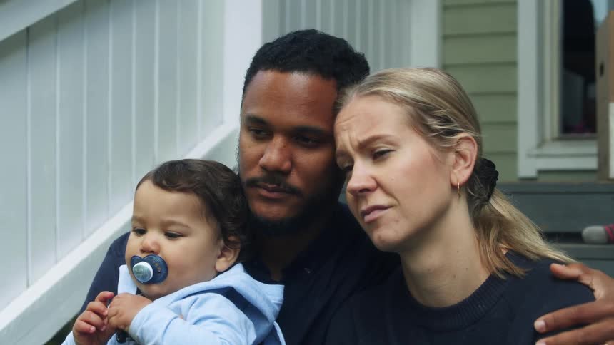 Man with beard black sweatshirt carrying toddler boy sits down next to blonde woman to console