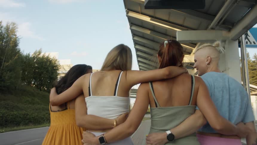 Four women walk together laughing with their arms around each others shoulders