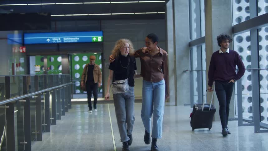 Two women in a terminal hugging and walking with a wall and windows in the background
