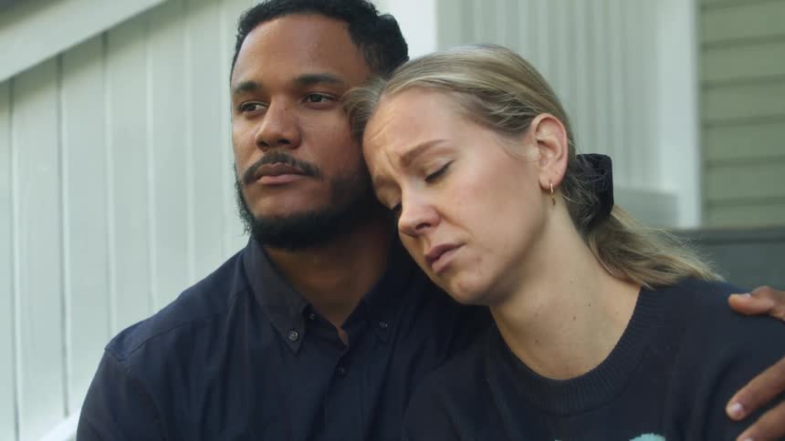 Man with black shirt consoles blonde woman with ponytail and black sweater sitting on stairs