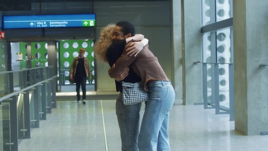 Woman hugs arriving friend in greeting as travelers come through doors at airport terminal