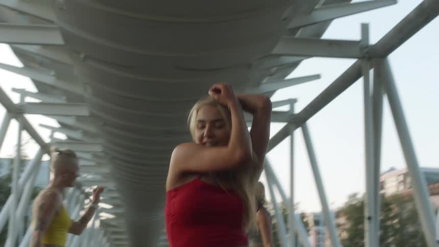 Four female dancers wearing cropped tops dance on pedestrian bridge with buildings in background