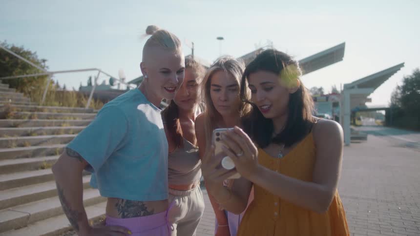 A group of women look at the screen of smartphone while standing together outdoors on a windy day