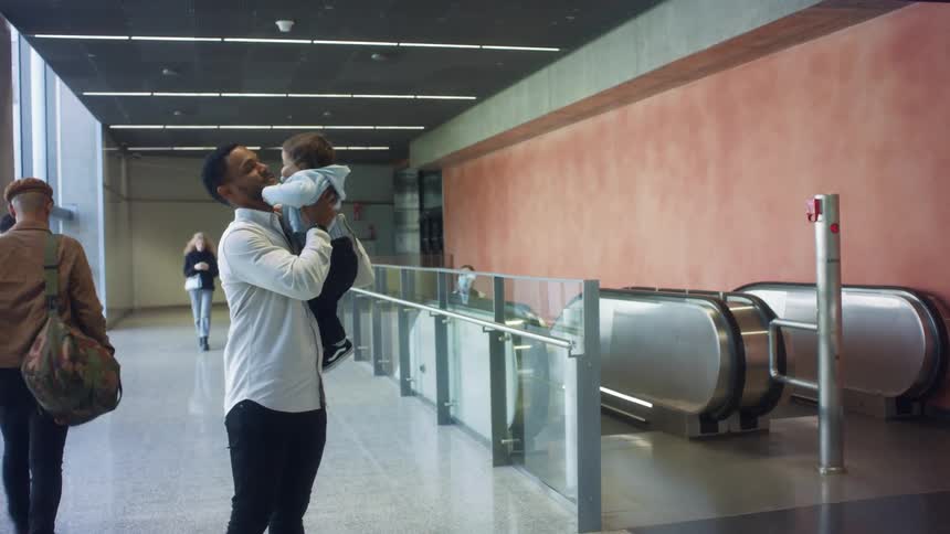 A man in a terminal holding a baby with an escalator and a wall in the background