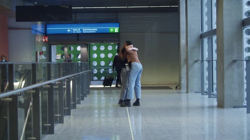 Woman waiting for friend greets her with hug as other travelers move along airport terminal