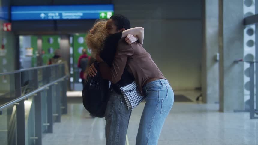 Woman with brown shirt and jeans and blonde woman with curly hair black shirt and jeans cuddle each other at the airport