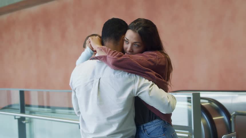 Man holding baby hugs woman in greeting as she walks off escalator with luggage