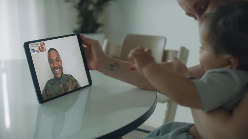 Baby toddler sits on mothers toddler making video call with man wearing a military uniform