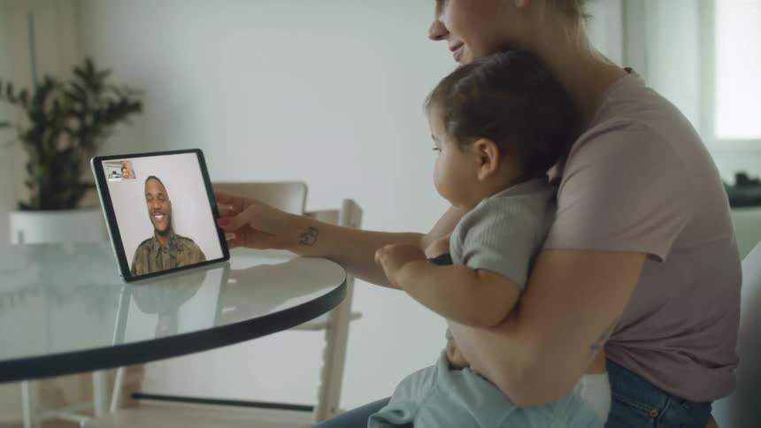 A woman holding a baby smiles as she holds a tablet on a video call with a man