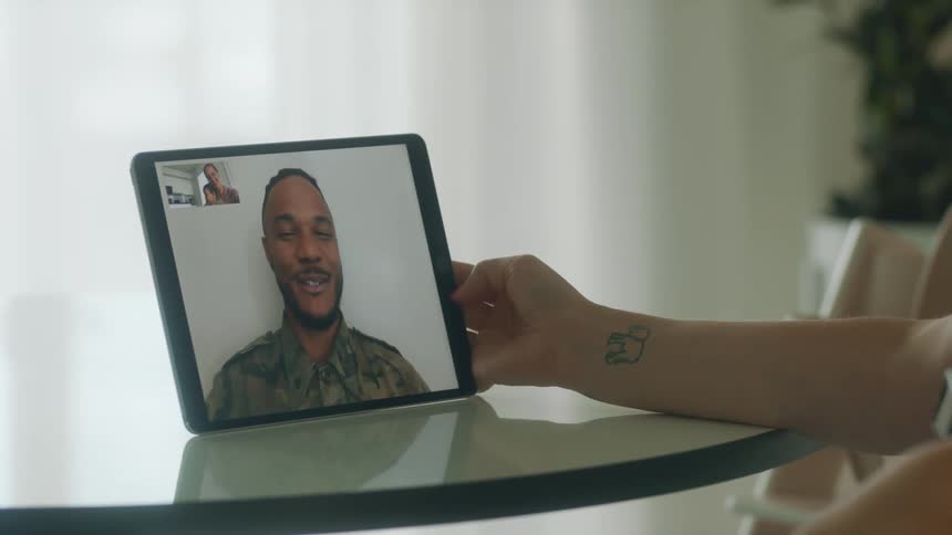 A woman holding a baby in her lap video chats with a man using a computer tablet while sitting at a table indoors